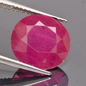 May include: A vibrant, oval-cut ruby gemstone with a deep pinkish-red hue and multifaceted surface, held securely by a pair of silver-toned metal tweezers. The gemstone displays internal clarity and light reflection across its polished facets.