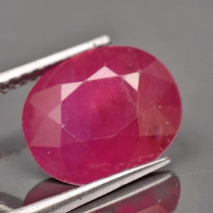 May include: A vibrant, oval-cut ruby gemstone with a faceted surface that reflects light. The deep pinkish-red stone is held by a pair of silver-toned metal tweezers, showcasing its translucent quality and precise geometric cut against a neutral background.