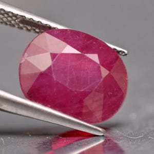 May include: A vibrant, oval-cut ruby gemstone with a deep pinkish-red hue and a faceted surface. The gemstone is held by a pair of silver-toned metal tweezers, showcasing its translucent quality and internal clarity characteristics under light.