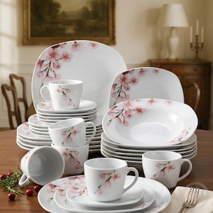 Puede incluir: Un juego de vajilla de porcelana blanca con un delicado patrón de flores de cerezo rosas. La colección incluye platos llanos cuadrados, platos de ensalada, cuencos para sopa y tazas a juego, decorados con suaves motivos florales en los bordes.