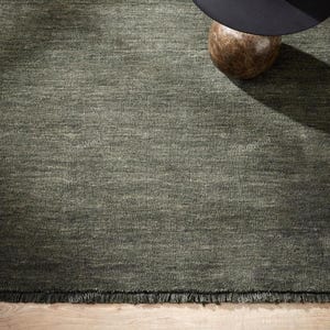 May include: A rectangular area rug in a deep olive green hue featuring a textured, woven surface with subtle horizontal variations in tone. The rug is finished with a short, fringed edge along the bottom and displays the text RugWind in white.