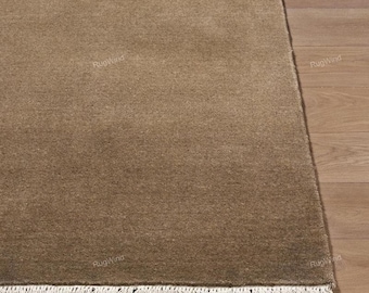 Op de afbeelding: Een rechthoekig vloerkleed in een effen, gedempte bruine tint met een dichte, laagpolige textuur. Het kleed heeft een rechte, strakke rand aan de bovenkant en zijkanten, afgewerkt met een rij korte, gebroken witte franjes aan de onderkant.