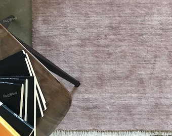 Op de afbeelding: Een oudroze vloerkleed met een getextureerd, geweven oppervlak en een korte, lichtgekleurde franje langs de onderrand. Het kleed ligt naast een donkerhouten tafeloppervlak met daarop verschillende zwarte boeken met de tekst RugWind erop.