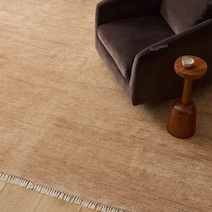 May include: A large, rectangular, light brown area rug with a textured surface and white fringe along the bottom edge. The rug features a subtle horizontal linear pattern and is positioned beneath a dark brown armchair and a small wooden side table.