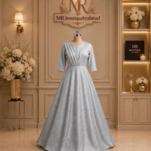 May include: A floor-length silver gown with a round neckline and three-quarter length sleeves. The bodice features vertical pleated detailing on the right side, transitioning into a full, flowing skirt with a subtle shimmering texture throughout.