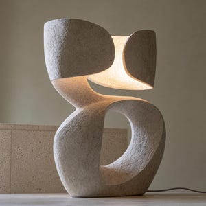 May include: A sculptural table lamp with a textured, beige stone-like finish. The lamp features an organic, curved form with a central open void and a top section that houses an integrated light source, casting a warm glow from within the structure.
