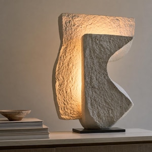 May include: A sculptural table lamp with a rough, stone-like off-white texture. The lamp features two interlocking, abstract geometric forms that create a warm, internal glow. It is mounted on a thin, rectangular black base.