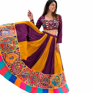 Op de afbeelding: Een tweedelige lehenga-set bestaande uit een paarse crop top met lange mouwen en spiegelwerk, en een veelkleurige rok. De rok heeft afwisselende paarse en gele panelen, een brede rand met cirkelvormige olifant- en pauwmotieven en een gestreepte zoom.