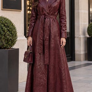 Può includere: Un trench lungo fino ai piedi, a maniche lunghe, di colore bordeaux intenso con un motivo testurizzato a pelle di serpente. Il cappotto presenta un ampio colletto a revers, una cintura da annodare in vita e una piccola borsa strutturata.