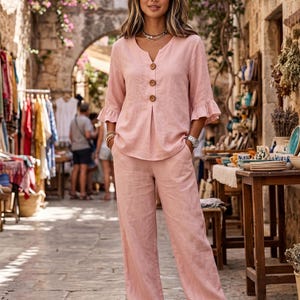 Puede incluir: Un conjunto de dos piezas en color rosa claro que consta de una blusa con cuello en V, tres botones marrones y mangas tres cuartos con volantes, combinado con pantalones anchos a juego. El conjunto es de tela texturizada y corte holgado.