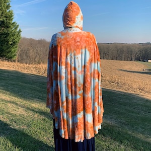 Puede incluir: Una capa con capucha con un estampado tie-dye en tonos naranja y azul claro. La capa se lleva sobre una falda azul oscuro. El fondo muestra un campo y árboles bajo un cielo azul.