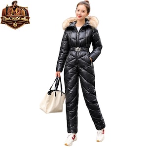 May include: A black quilted puffer jumpsuit featuring a high collar with a faux fur hood, a front zip closure, and a belted waist with a silver buckle. The garment has long sleeves and is paired with a large cream-coloured tote bag.