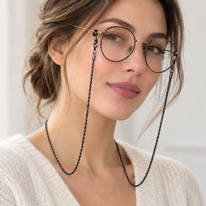 Op de afbeelding: Een bril met een rond montuur, dunne goudkleurige metalen randen en heldere glazen, bevestigd aan een lange, slanke zwarte metalen ketting. De ketting heeft kleine, uniforme schakels en is met zwarte rubberen lusjes aan de pootjes bevestigd.