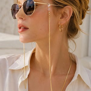 Op de afbeelding: Een goudkleurige metalen ketting voor brillen met een fijn schakelontwerp. De ketting wordt met doorzichtige siliconen lusjes aan de pootjes van de zonnebril bevestigd, waardoor deze in een lange, slanke lijn langs de zijkanten hangt.