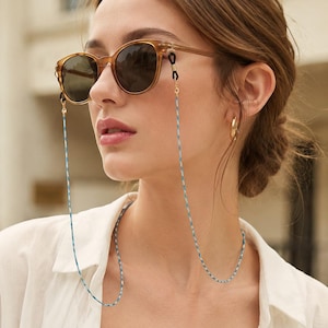 May include: Brown tortoiseshell sunglasses with dark lenses attached to a long, thin, braided cord. The cord features a repeating pattern of light blue and white segments, connected to the frames by small black rubber loops and gold metal hardware.