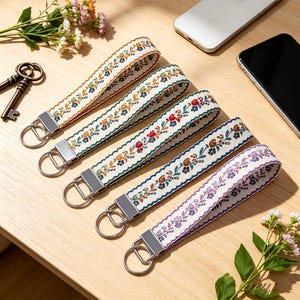 Könnte beinhalten: Fünf Stoff-Schlüsselanhänger mit silberfarbenen Metallbeschlägen und Schlüsselringen. Jedes Band zeigt ein einzigartiges gesticktes Blumenmuster in verschiedenen Farben wie Blau, Orange, Rot, Braun und Violett auf cremefarbenem Grund.