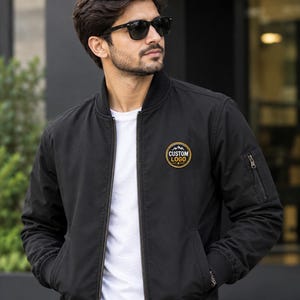 Peut inclure: Un blouson bomber noir porté sur un t-shirt blanc. La veste présente une fermeture éclair frontale, un col côtelé et un écusson circulaire sur la poitrine gauche avec le texte CUSTOM LOGO et un motif de montagne. Une poche zippée est sur la manche.
