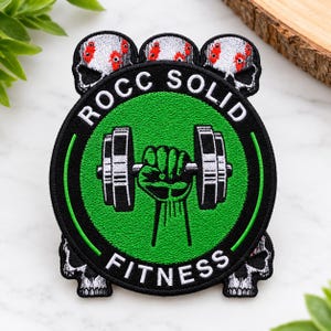 May include: A circular embroidered patch featuring a green center with a black fist holding a dumbbell. The black border contains the white text ROCC SOLID FITNESS. Three white skulls with red accents are positioned at the top and two at the bottom.