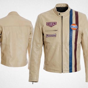 Può includere: Una giacca in pelle beige con colletto alla coreana, cerniera frontale e due tasche laterali. La parte anteriore presenta una striscia verticale blu e bordeaux, un logo circolare Gulf e toppe rettangolari con il testo Heuer e Firestone.