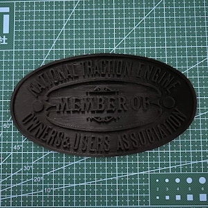 Può includere: Una targa ovale nera con scritte in rilievo che recita NATIONAL TRACTION ENGINE MEMBER OF OWNERS & USERS ASSOCIATION. La targa è posizionata su un tappetino da taglio verde con una griglia e segni in centimetri e gradi per riferimento.