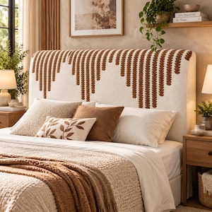 May include: A cream-coloured upholstered headboard featuring a repeating vertical pattern of embroidered rust-coloured leaves arranged in a scalloped design. The headboard is positioned behind a bed with white sheets, a textured cream blanket, and pillows.