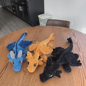 May include: Three plush dragon toys made of soft, textured yarn. One is blue with light gray horns, one is golden-orange, and one is black with a gray tail tip. Each dragon features large, round, glossy eyes and detailed wings and limbs.