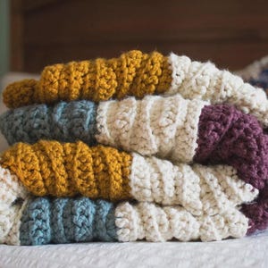 Pode incluir: Uma manta grossa feita de crochê com um padrão de blocos de cores amarelo mostarda, creme, azul suave e roxo ameixa profundo. O fio apresenta um ponto grosso e texturizado, criando uma aparência macia e dobrada com listras horizontais.