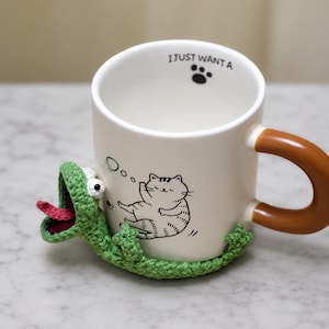 May include: A white ceramic mug with a brown handle featuring a black line drawing of a cat and the text I JUST WANT A with a paw print inside the rim. A green crocheted lizard with a red tongue is attached to the base of the mug.