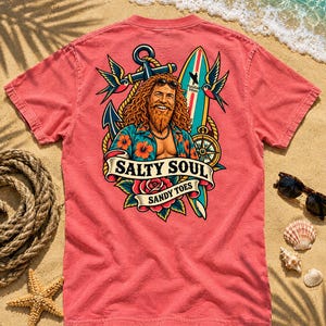 May include: A coral-colored short-sleeve t-shirt featuring a graphic on the back. The design depicts a bearded person with long hair wearing sunglasses and a floral shirt, surrounded by an anchor, surfboard, compass, swallows, and roses, with the text SALTY SOUL SANDY TOES.