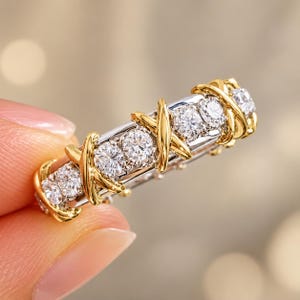 Puede incluir: Un anillo bicolor con una banda de color plateado engastada con una serie de piedras redondas, transparentes y facetadas. La banda está acentuada con superposiciones metálicas en forma de X de color oro amarillo pulido.