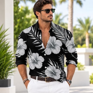 May include: A long-sleeved button-down shirt featuring a black background with a repeating pattern of large white hibiscus flowers and palm leaves. The shirt has a classic collar, white buttons down the front, and cuffed sleeves.
