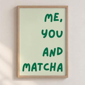 May include: A rectangular wall art print in a light wooden frame featuring a pale green background with the dark green text "ME, YOU AND MATCHA" written in a rounded, bold, sans-serif font centred vertically on the print surface.