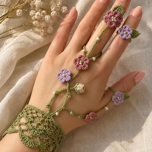 May include: A green crochet hand flower bracelet featuring delicate floral motifs in shades of mauve and lavender. The design includes intricate green vine-like strands adorned with small white beads, connecting a wide wrist cuff to two rings.