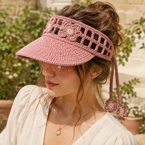 May include: A dusty rose crochet sun visor featuring a wide brim and a decorative band with openwork grid patterns and floral motifs. Two long crochet ties hang from the sides, each ending in a small flower accent with a pale yellow center.