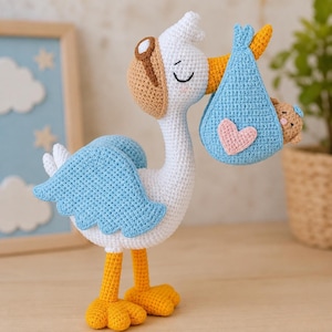 Pode incluir: Um brinquedo de cegonha em crochê branco e azul claro com pés e bico amarelos, usando óculos de aviador castanhos. A cegonha carrega um embrulho azul em crochê com um coração rosa, contendo uma pequena cabeça de urso castanha com um laço azul.