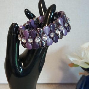 Op de afbeelding: Een elastische armband met meerdere rijen bloemvormige kralen in verschillende tinten licht- en donkerpaars, afgewisseld met heldere, ronde, gefacetteerde strass-steentjes in zilverkleurige metalen zettingen door het ontwerp.