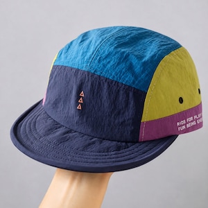 Könnte beinhalten: Eine Fünf-Panel-Kappe mit einem Color-Block-Design in Blau, Marineblau, Olivgrün und Lila. Das vordere marineblaue Panel zeigt drei kleine orangefarbene Dreiecke, während das lila Seitenpanel den weißen Text RIDE FOR PLAY FUN BEING ENS trägt.