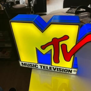Peut inclure: Une enseigne lumineuse rectangulaire représentant le logo MTV avec un grand M jaune, un V bleu et l'inscription TV en rouge. La base est blanche avec le texte noir MUSIC TELEVISION et un symbole de marque déposée. L'enseigne émet une lueur vive.