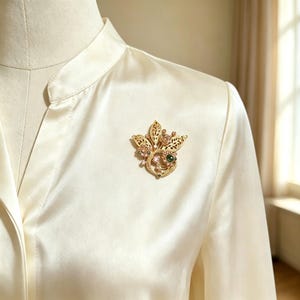 Puede incluir: Un broche de metal dorado con forma de ramo floral con hojas de filigrana, pequeños cristales rosas y una piedra cabujón verde oscuro, prendido en una blusa de satén color crema con cuello mao y tapeta de botones frontal.