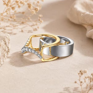 Puede incluir: Un conjunto de dos anillos compuesto por una banda ancha de tono plateado pulido y un anillo de marco abierto en tono dorado. El anillo dorado presenta una sección curva adornada con una hilera de pequeñas piedras claras y brillantes.