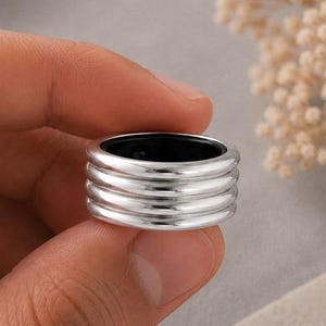 Peut inclure: Une bague large de couleur argentée présentant un design empilé avec quatre bandes horizontales distinctes. La surface extérieure a une finition métallique polie et réfléchissante, tandis que l'intérieur est doublé d'un matériau noir lisse.