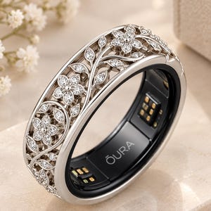 Puede incluir: Un anillo inteligente de tono plateado con un intrincado diseño floral calado y piedras transparentes incrustadas. La banda exterior muestra un patrón de enredaderas y flores, mientras que la banda interior es negra con el texto OURA y sensores.