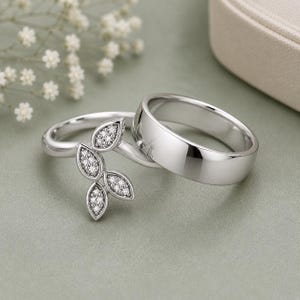 Puede incluir: Un conjunto de dos anillos de tono plateado. Un anillo presenta una banda lisa y pulida. El segundo anillo tiene una banda delicada adornada con un grupo de cuatro elementos en forma de hoja, cada uno incrustado con pequeñas piedras claras.