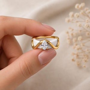 Peut inclure: Une bague dorée composée d'un anneau blanc lisse avec une superposition dorée ondulée. Une pierre précieuse transparente en forme de poire est sertie au centre de l'ondulation, créant un point focal. Le design associe métal doré et base blanche.