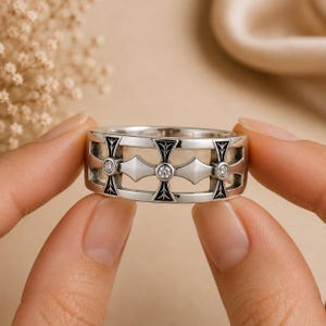 Puede incluir: Un anillo de banda en tono plateado con un patrón calado repetitivo de motivos en forma de cruz. Cada cruz está acentuada con una pequeña piedra preciosa redonda y transparente en el centro y detalles de esmalte negro en los brazos.