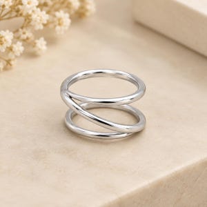 Peut inclure: Une bague argentée polie présentant un design multicouche avec deux anneaux distincts qui se chevauchent dans un mouvement continu en spirale, créant une structure ouverte et aérée avec une finition métallique lisse et réfléchissante.