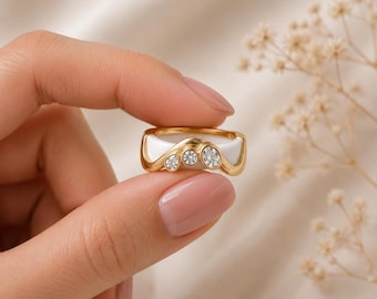 May include: A gold-toned ring featuring a smooth white band inlay and a curved top section set with three clear, round-cut stones. The metal frame follows a wave-like contour, creating a layered appearance with the white material.