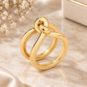 Puede incluir: Un anillo de tono dorado pulido que presenta una banda lisa y continua que se entrelaza en la parte superior para crear un diseño de nudo prominente. La superficie metálica es altamente reflectante y muestra un tono amarillo cálido y metálico.