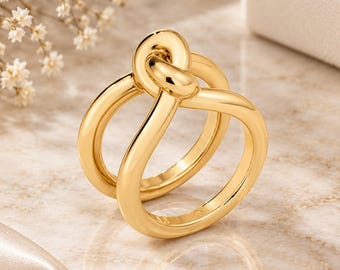 May include: A polished gold-tone ring featuring a continuous, smooth band that loops over itself at the top to create a prominent knot design. The metal surface is highly reflective, showcasing a warm, metallic yellow hue throughout the entire piece.