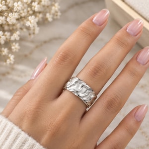 May include: A wide silver ring featuring a textured, wavy pattern across the band. The surface of the ring has a matte, granular finish within the recessed areas, while the raised edges of the wave design are polished to a bright, reflective shine.